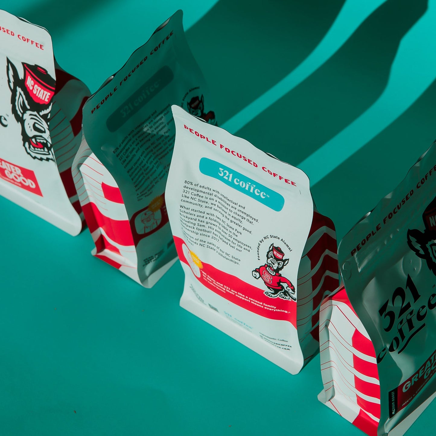 Four bags of Greater Good coffee are placed next to each other on a teal background. Each bag is rotated 90 degrees, creating a line up of bags that display all four sides of the packaging.