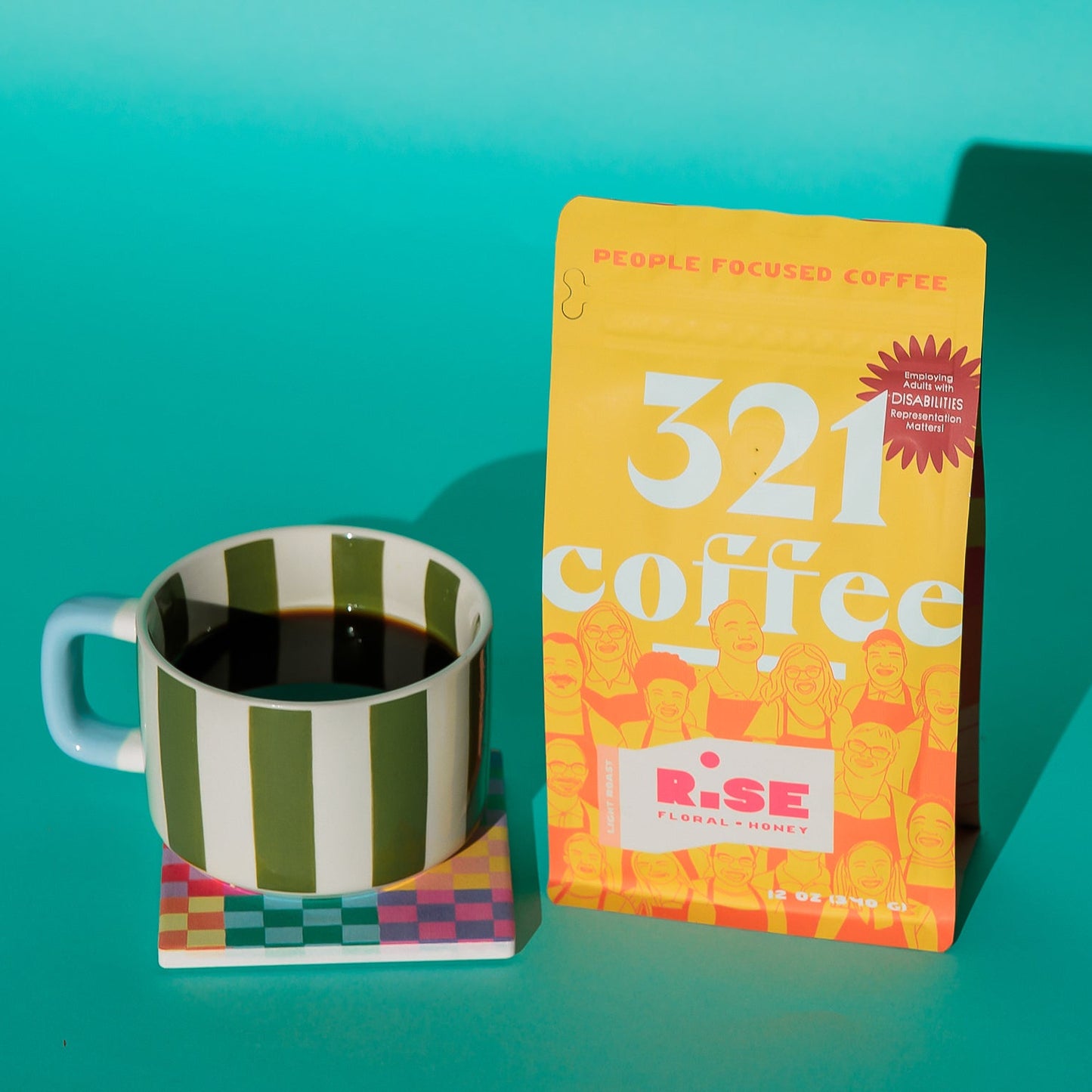 A yellow 12 ounce bag of Rise, 321 Coffee's light roast coffee, sits next to a green and white striped ceramic mug that is full of hot coffee. The mug rests on a colorful checker print coast, and the whole scene sits on a teal background.