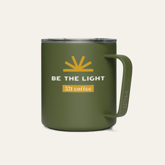 A dark green metal camp mug with "Be the Light" printed in white on the mug with a yellow sun above it.