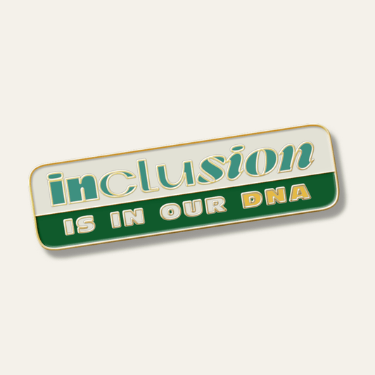An enamel pin in the shape of a rectangle with "inlcusion" written in green on a tap background above "is in our DNA" written in tan and yellow on a green background.