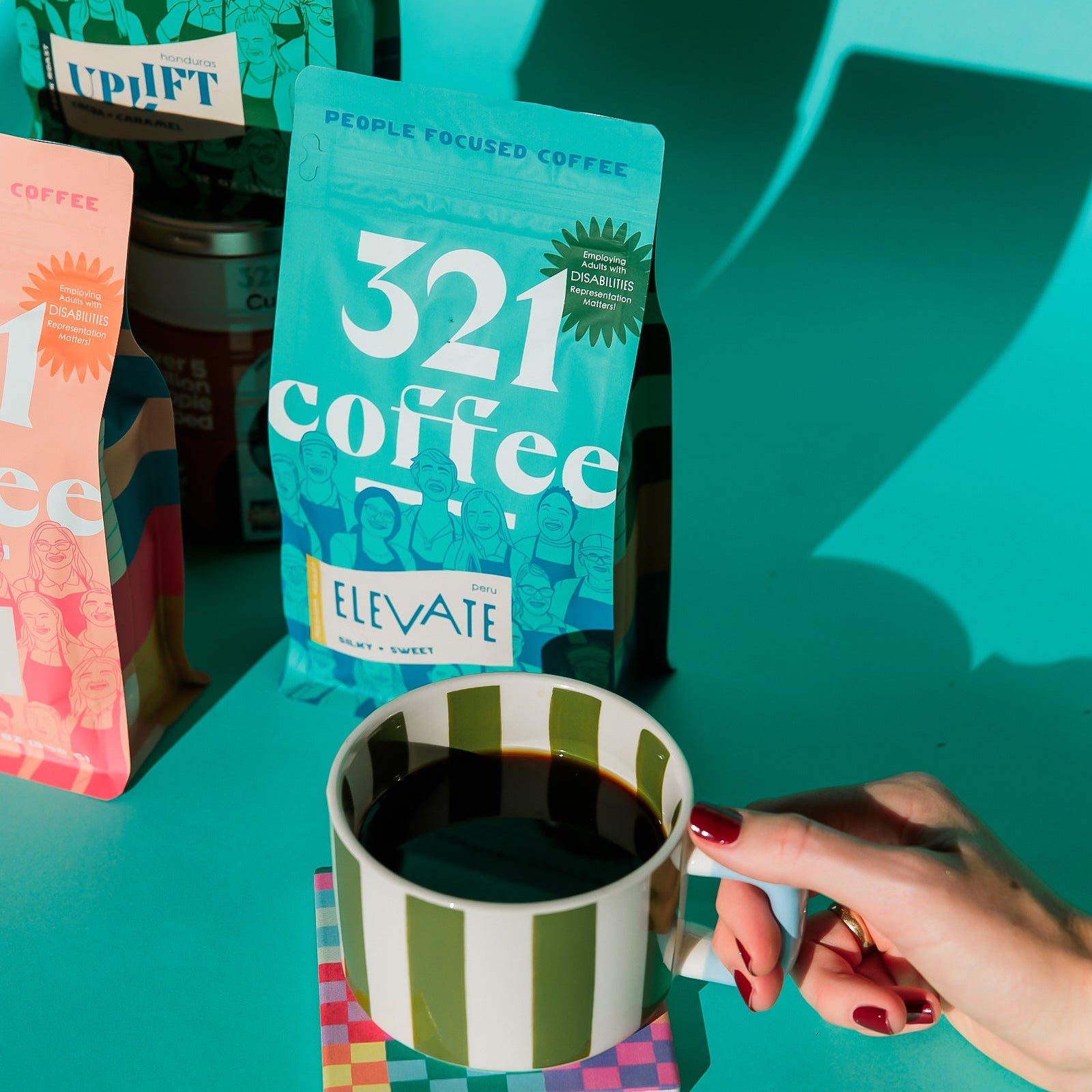 A teal 12 ounce bag of Elevate, 321 Coffee's medium roast coffee, rests on a teal background, placed behind a white and green striped ceramic mug. A hand reaches out from the bottom right corner to hold the handle of the mug, as if just about to pick up the mug.