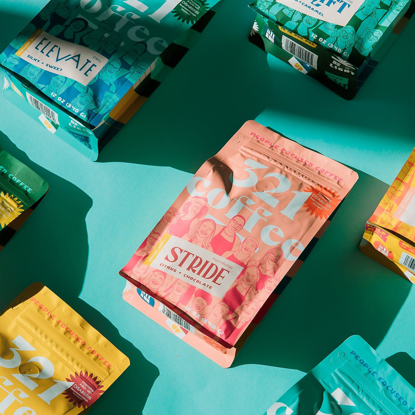 A group of 12 oz bags of 321 Coffee all lay flat on a teal background. All the bags are angled diagonally up and to the right. A pink bag of Stride, 321 Coffee's decaf coffee, is in the center of the display.