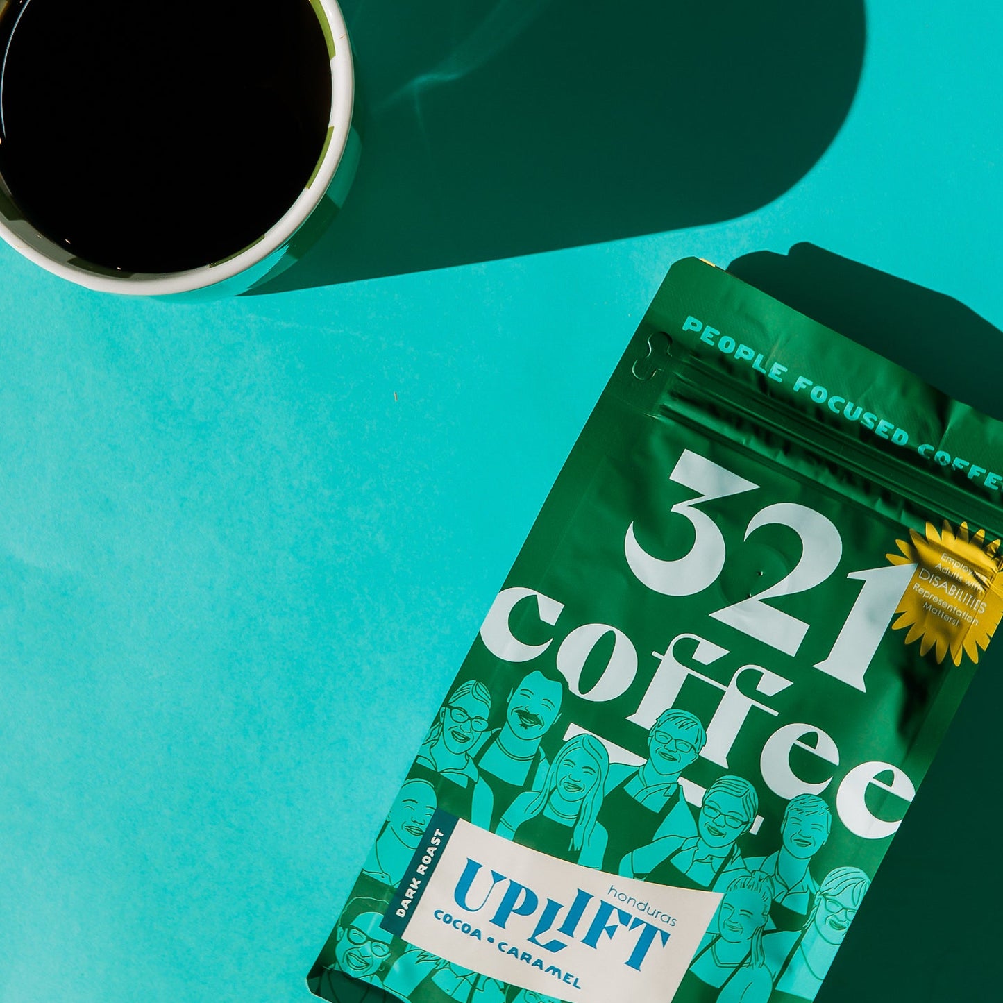 A 12 ounce green bag of Uplift, 321 Coffee's dark roast coffee, lays flat on a teal background. A mug of hot coffee rests to the top left corner of the bag.