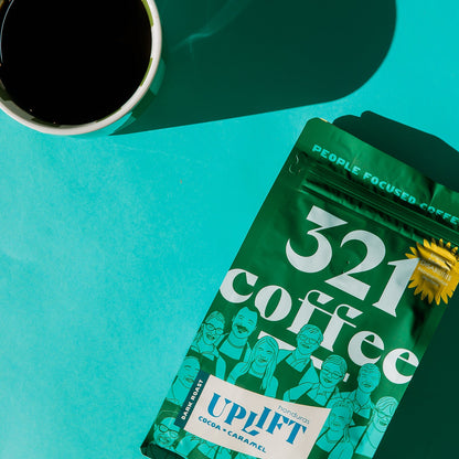 A 12 ounce green bag of Uplift, 321 Coffee's dark roast coffee, lays flat on a teal background. A mug of hot coffee rests to the top left corner of the bag.
